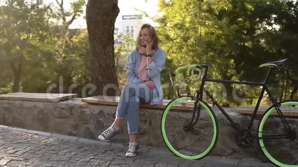 漂亮的女孩坐在城市公园的长凳或栏杆上旁边有她的徒步旅行自行车她的手机在说话视频的预览图