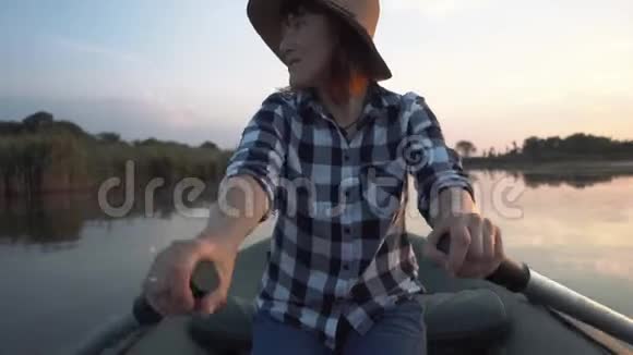 戴着帽子的年轻快乐女孩在湖上用木桨的充气船上航行视频的预览图