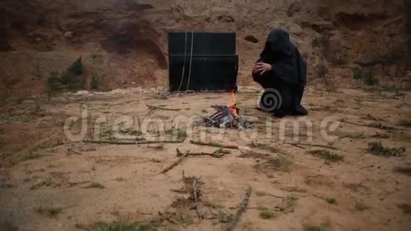 一个穿着黑袍带着头巾的和尚坐在空地上在炉火旁取暖视频的预览图