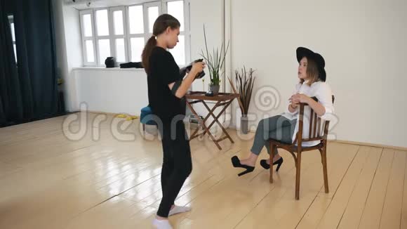 为身穿黑色衣服的年轻女摄影师做特写向身穿黑色帽子白色衬衫的时尚年轻模特展示照片视频的预览图