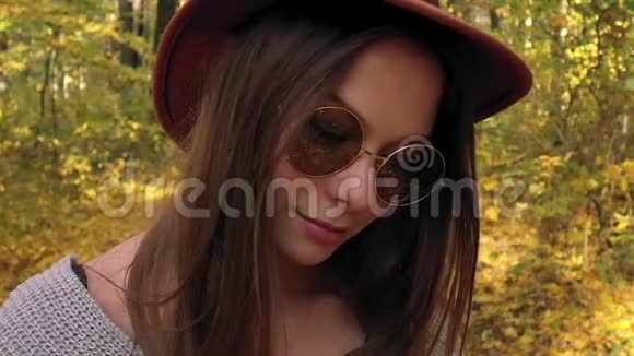 一位戴着帽子戴着墨镜戴着黄色枫叶的美丽微笑女孩在秋天的前景中的肖像视频的预览图