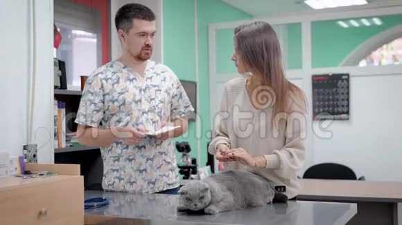 一位成年男性兽医正在办公室咨询一位年轻女性女孩正抚摸躺在桌子上的灰猫视频的预览图