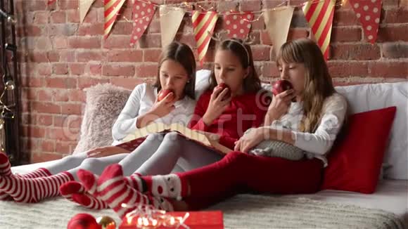 三姐妹穿着圣诞睡衣坐在床上吃着红苹果三胞胎女孩读了一本圣诞故事书视频的预览图