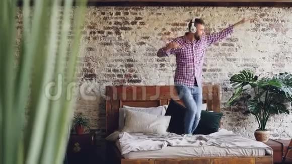 穿着牛仔裤和衬衫的留着胡子的人正在双人床上跳舞一边唱歌一边用无线方式听音乐视频的预览图