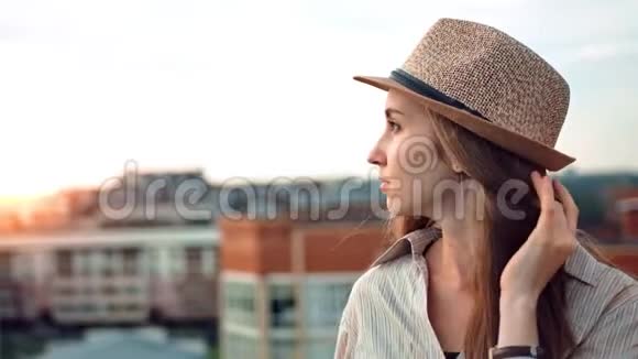 侧观忧郁的女人戴着帽子在屋顶或阳台上令人惊叹的日落中欣赏城市景观视频的预览图