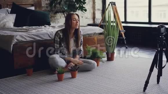 一位著名的年轻女性博客作者在双人床附近的卧室里坐着一边用摄像机录制关于盆花的视频视频的预览图