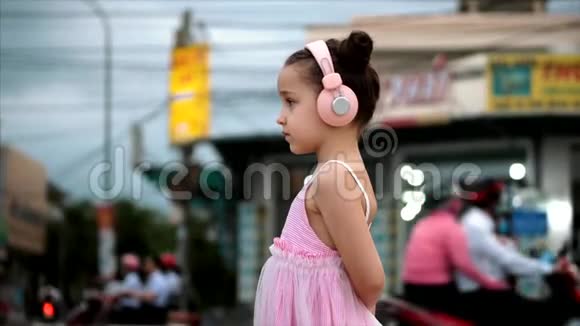 一个可爱的孩子站在路边的肖像一个可爱的小女孩穿着粉红色的连衣裙和粉红色的耳机看着视频的预览图