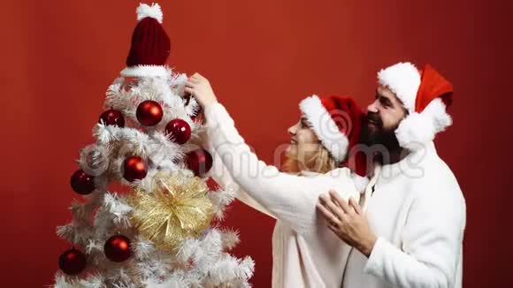可爱的年轻夫妇在红色的背景上装饰一棵圣诞树大胡子男人拥抱装饰圣诞树的女人视频的预览图
