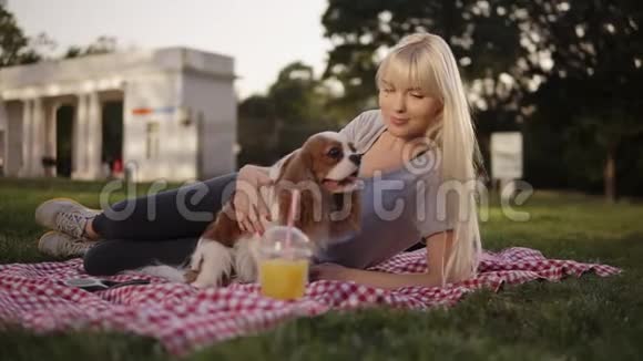 金发碧眼的女人躺在公园的一块地上躺在一个格子的垃圾上抚摸着她的小狗背景模糊视频的预览图