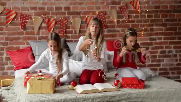 三个穿着圣诞睡衣的姐妹坐在装饰好的床上过圣诞节三胞胎女孩打开礼物视频的预览图