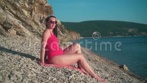 美丽的怀孕女孩穿着红色的一件泳衣和眼镜坐在海滩上视频的预览图