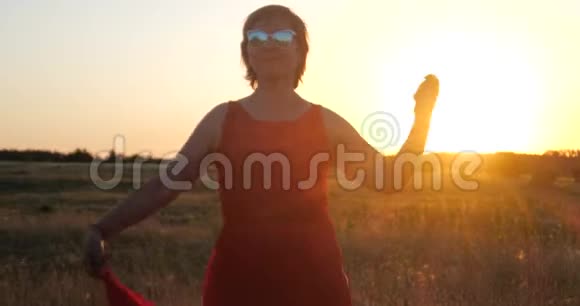 在夕阳下穿着红色裙子在大片田野里跳舞的时髦女人微笑着视频的预览图