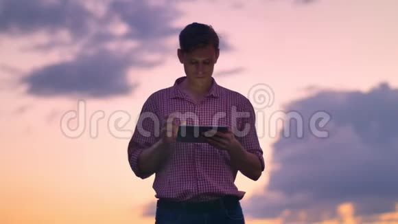 英俊自信的年轻人穿着衬衫在平板电脑上打字向前看在粉红色的天空上孤立背景是夕阳视频的预览图