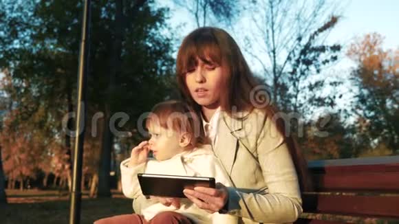 妈妈和孩子坐在公园的长凳上看书在平板电脑上视频的预览图