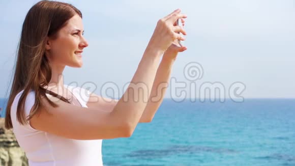 微笑的年轻女子在手机上慢动作拍摄岩石和蓝色地中海视频的预览图