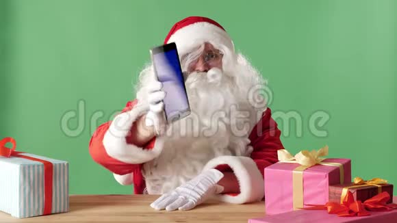 笑容可掬的圣诞老人展示平板电脑相机里有蓝色屏幕桌子上有礼物背景是彩色的视频的预览图
