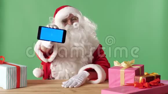 微笑圣诞老人在镜头中展示智能手机礼物在桌子上背景中的色度视频的预览图