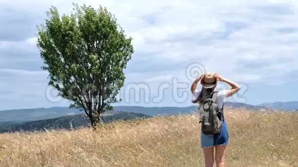 带帽子和背包的年轻女子在田野里的后景草地干草环绕着群山视频的预览图