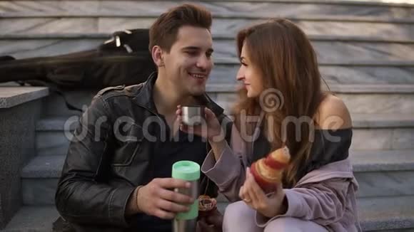 年轻夫妇坐在楼梯上吃羊角面包在户外喝咖啡坐在格子布上说话视频的预览图