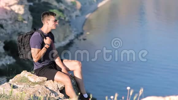 侧观英俊的欧洲年轻男性旅行者坐在悬崖附近干净的池塘中长镜头视频的预览图