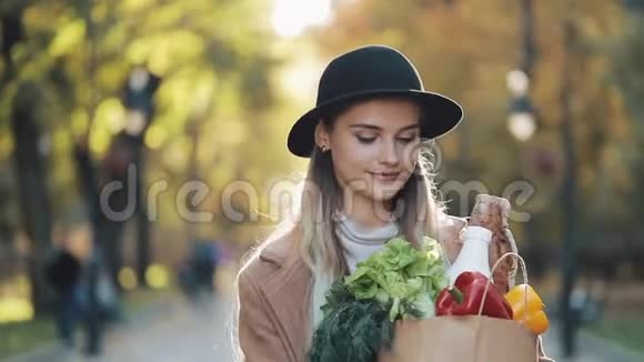 年轻漂亮的女人穿着时髦的外套在公园里散步拿着一包产品微笑着环顾四周视频的预览图