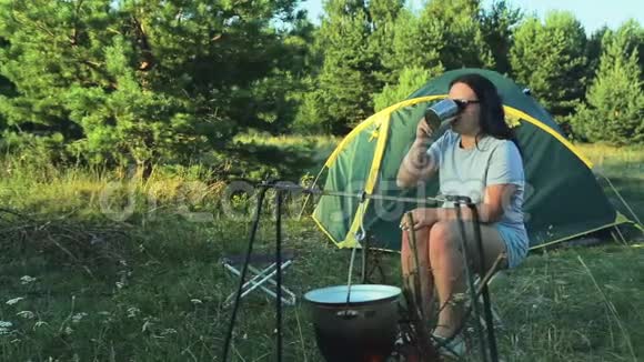 一男一女戴着墨镜坐在帐篷旁喝着茶看着锅子挂在火堆上视频的预览图