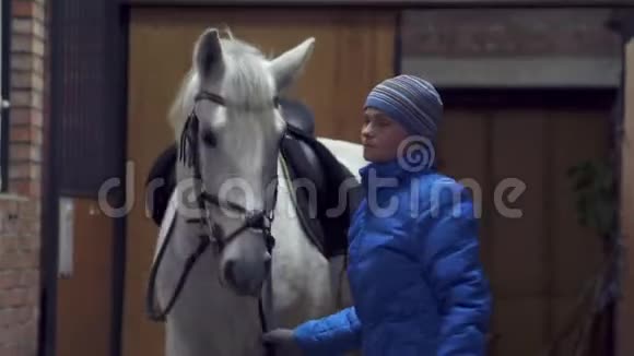 一个穿着蓝色夹克和运动帽的年轻漂亮女人把一匹白马从马厩里带出来骑马视频的预览图