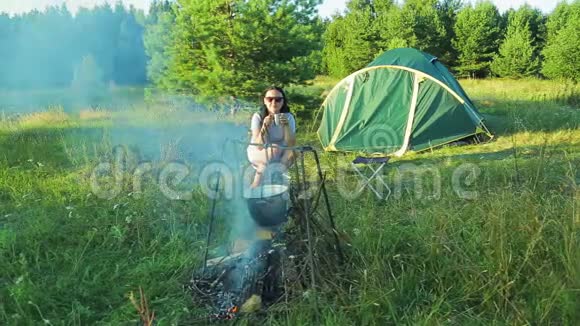 一个年轻的女人坐在帐篷附近的火旁看着火保龄球在火上挂着喝着茶视频的预览图