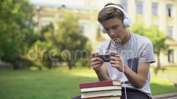 十几岁的男孩戴着耳机玩手机游戏代替作业拖拖拉拉视频的预览图