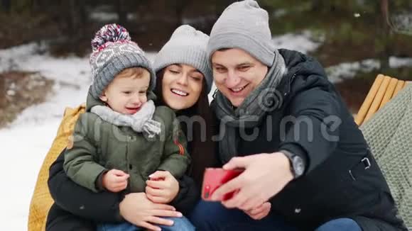 家人和儿子在电话里自拍他们坐在冬天森林里的沙发上篝火覆盖葡萄酒圣诞快乐视频的预览图