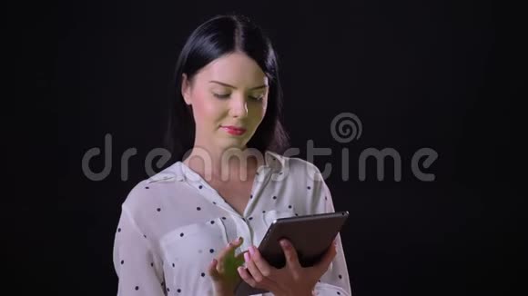 年轻的黑发女人带着迷人的色彩和微笑的平板电脑站在黑色的背景上显得很迷人视频的预览图