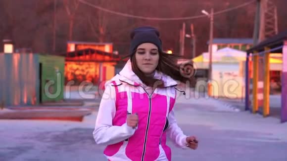 慢动作年轻苗条的女孩戴着运动帽和运动夹克早上在水泥路上跑步视频的预览图