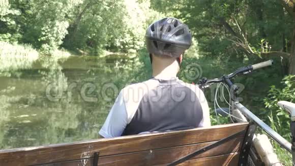 一个骑自行车的人夏天在公园里休息他坐在池塘附近的长凳上他用音乐听音乐视频的预览图