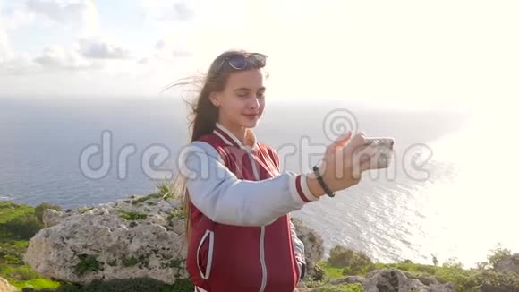 金发女孩在大自然岩石海洋绿色植物的背景下自拍长发少女微笑着视频的预览图