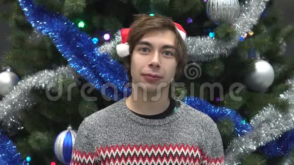 圣诞老人帽子里英俊的年轻人的肖像看圣诞树背景上的相机视频的预览图