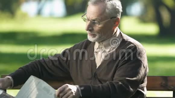 戴眼镜看报纸的退休老人早上坐在公园的长椅上视频的预览图