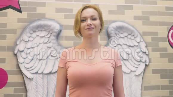 一个金发女人站在墙的背景上以天使的翅膀的形式展开视频的预览图