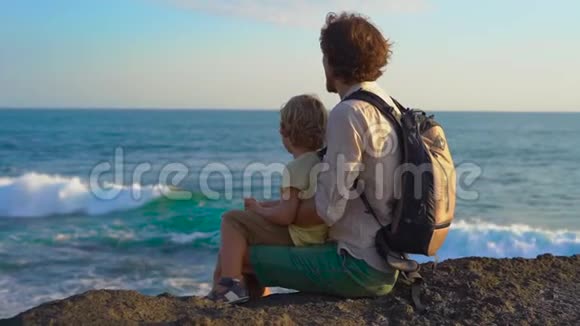 一对父子坐在岩石上看着塔纳罗得寺附近的海浪慢镜头视频的预览图