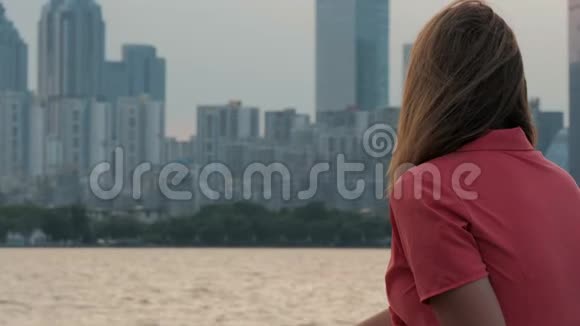 穿着粉红色裙子的年轻女子欣赏日落时分坐在海滩上的城市风景视频的预览图