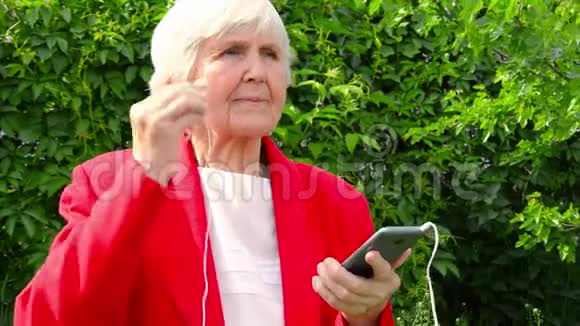老太太奶奶震惊了在太阳日的户外现代白种人奶奶穿着红色夹克听着耳机里的音乐视频的预览图