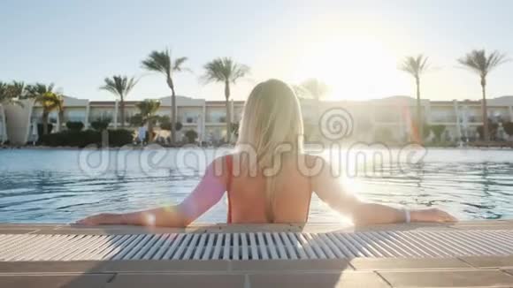 后景肖像金发少女看日落穿红色泳衣的女人坐在蓝色的游泳池里比基尼视频的预览图