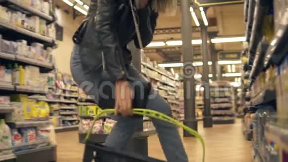 穿牛仔裤和黑色皮夹克的金发女人蹲着从下层货架上挑选产品然后把它放在上面视频的预览图