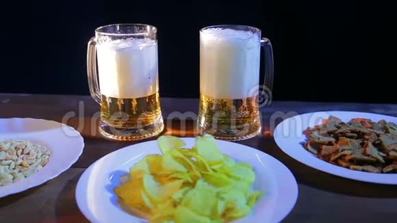 在黑色背景的酒吧里的木桌上放着两个起泡啤酒的杯子在盘子附近有薯片和坚果视频的预览图