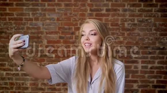 年轻的白种人金发女郎自拍微笑照片智能手机肖像砖墙的背景视频的预览图