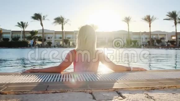 后景肖像金发少女看日落穿红色泳衣的女人坐在蓝色的游泳池里比基尼视频的预览图