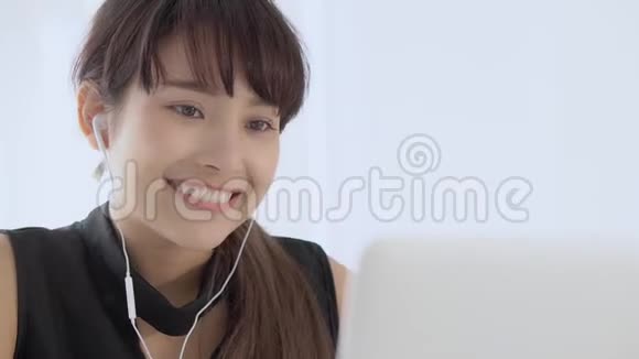 美丽的年轻亚洲女人微笑着坐在办公室里用手提电脑听带耳机的音乐放松视频的预览图