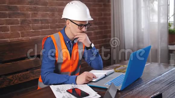 建筑公司的工程师穿着工作服和防护头盔在房间里的笔记本电脑上工作视频的预览图