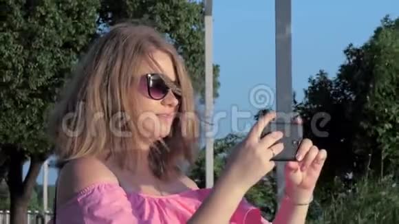 戴墨镜的美丽快乐的白种人年轻女子的肖像智能手机拍摄美丽的日落视频的预览图