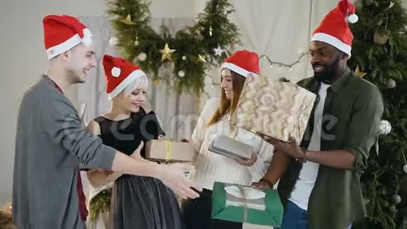戴圣诞帽的年轻快乐的朋友站在圣诞树前手里拿着礼物盒和圣诞礼物视频的预览图