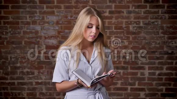 年轻的白种人金发女郎微笑着看书看着镜头肖像背景中的砖墙视频的预览图